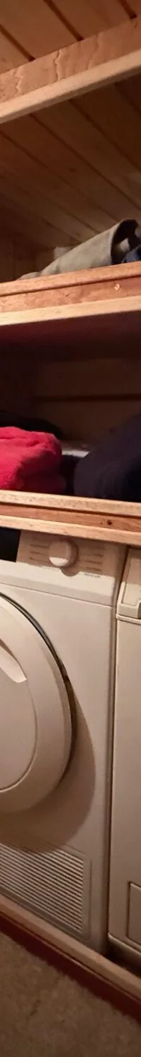 Een wasmachine en droger staan onder houten planken met opgevouwen kleding en een tas, in een houten ruimte.