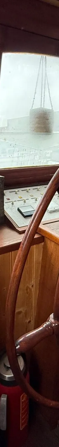 De foto toont het interieur van een stuurhut met een groot houten stuurwiel, diverse instrumenten en ramen met uitzicht…