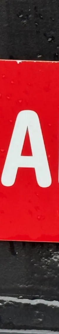 Een rode, langwerpige plaat met afgeronde hoeken en de witte tekst "JAN ABELS" is vastgeschroefd op een donkere ondergrond.