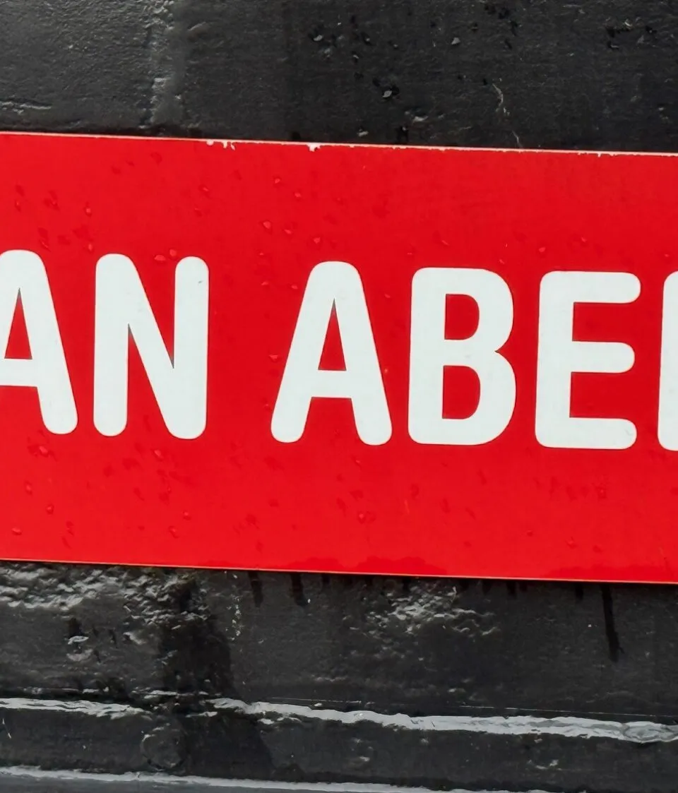 Een rode, langwerpige plaat met afgeronde hoeken en de witte tekst "JAN ABELS" is vastgeschroefd op een donkere ondergrond.