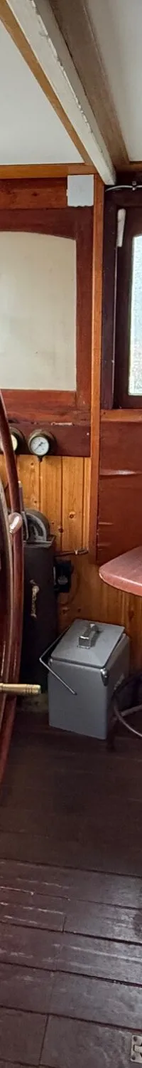 De foto toont een houten stuurhut met een groot houten stuurwiel, diverse ramen en een kruk.