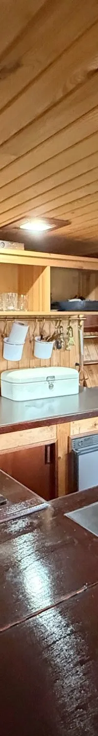 De foto toont een houten scheepskeuken met een fornuis, aanrecht, opbergruimte en een hangende koperen lamp.