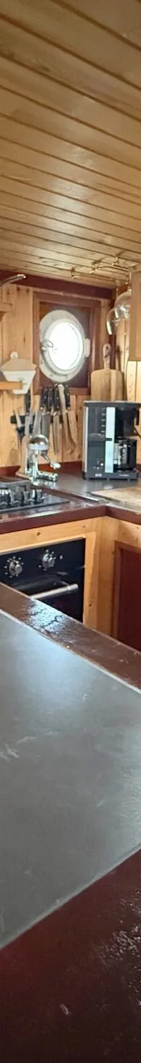 Een houten scheepskeuken met een kookplaat, spoelbak, koelkast, opbergplanken en een koperen lamp.