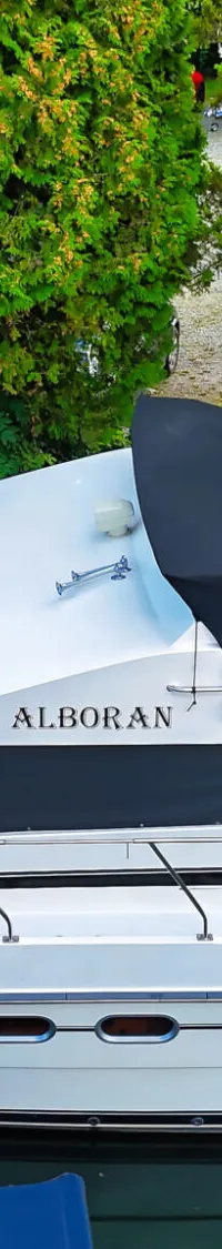 Een witte motorboot met de naam "ALBORAN" ligt in het water, omringd door groen en met mensen op de achtergrond.