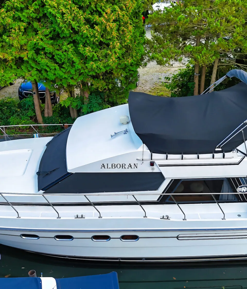 Een witte motorboot met de naam "ALBORAN" ligt in het water, omringd door groen en met mensen op de achtergrond.