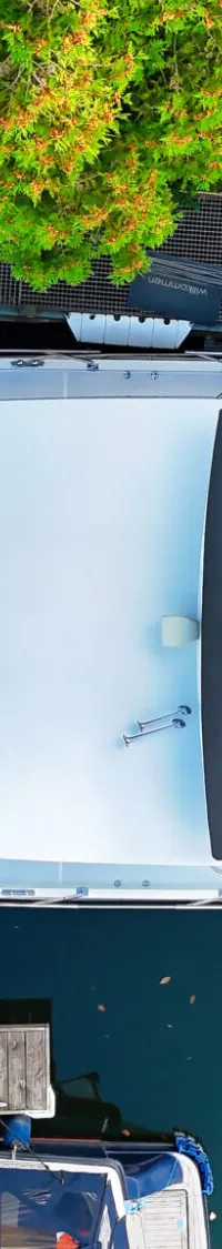 Een witte boot met een zwart dekzeil ligt aangemeerd aan een houten steiger, omringd door groen.