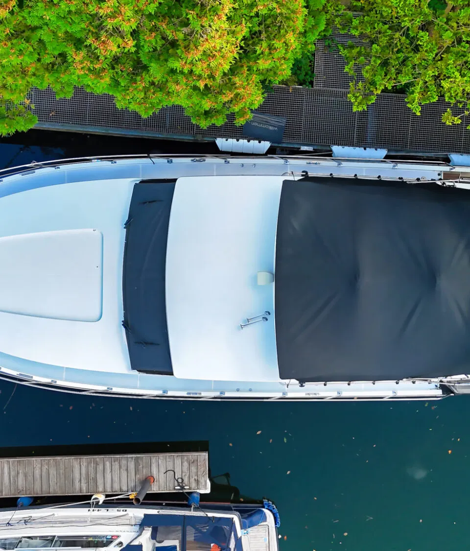 Een witte boot met een zwart dekzeil ligt aangemeerd aan een houten steiger, omringd door groen.