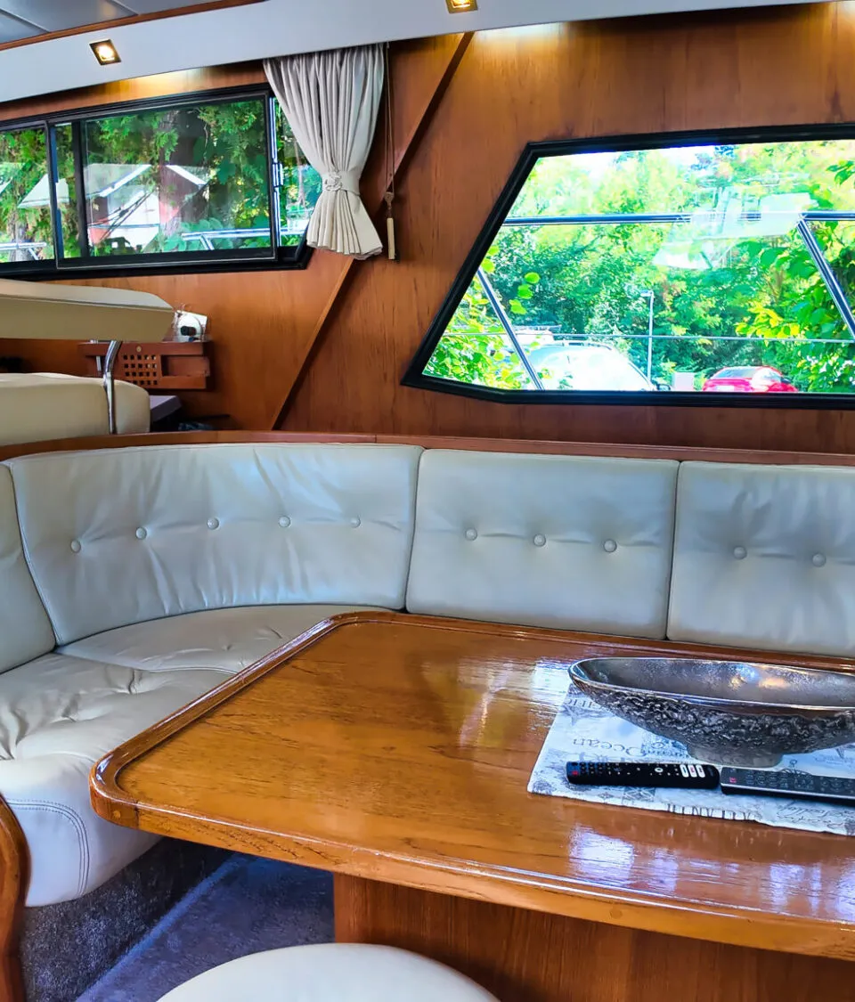 Een interieur van een boot met een houten tafel, een crèmekleurige bank en een stuurstand met een stuurwiel.