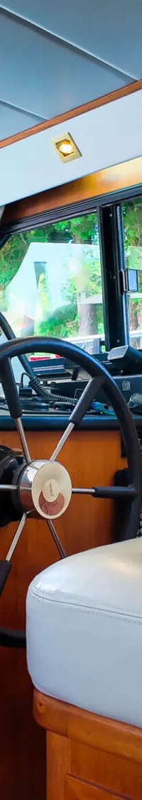 De foto toont een stuurstand van een boot met een stuurwiel, instrumentenpaneel, een witte stoel en ramen met uitzicht op…