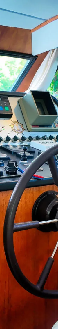 De foto toont het interieur van een stuurhut met een stuurwiel, navigatieapparatuur en houten panelen.