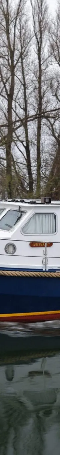 Een blauw-witte motorboot met de naam "BUTSKOP" vaart op het water, met kale bomen op de achtergrond.
