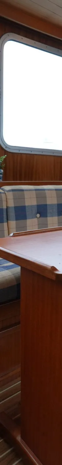 De foto toont een interieur van een boot met een houten tafel, een bank met blauw-beige ruitpatroon kussens en houten wanden.