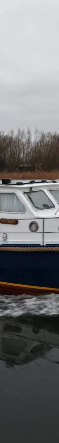 Een blauw-witte motorboot met een Nederlandse vlag vaart over water met een bewolkte lucht en bomen op de achtergrond.