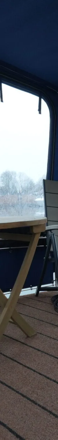 Een overdekt buitendek met twee klapstoelen, een klaptafel en een bruine vloerbedekking met zwarte strepen.