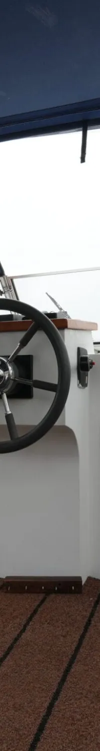 De foto toont de stuurstand van een boot met een donkerblauw dekzeil, een stuurwiel en een bruine vloer.