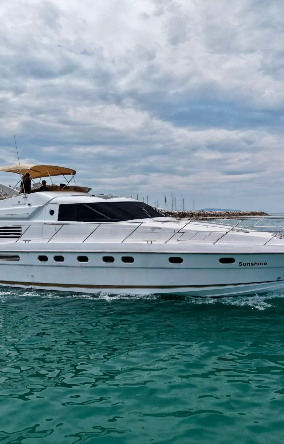 Een witte motorjacht met de naam "Sunshine" vaart op zee onder een bewolkte hemel, met een kustlijn op de achtergrond.