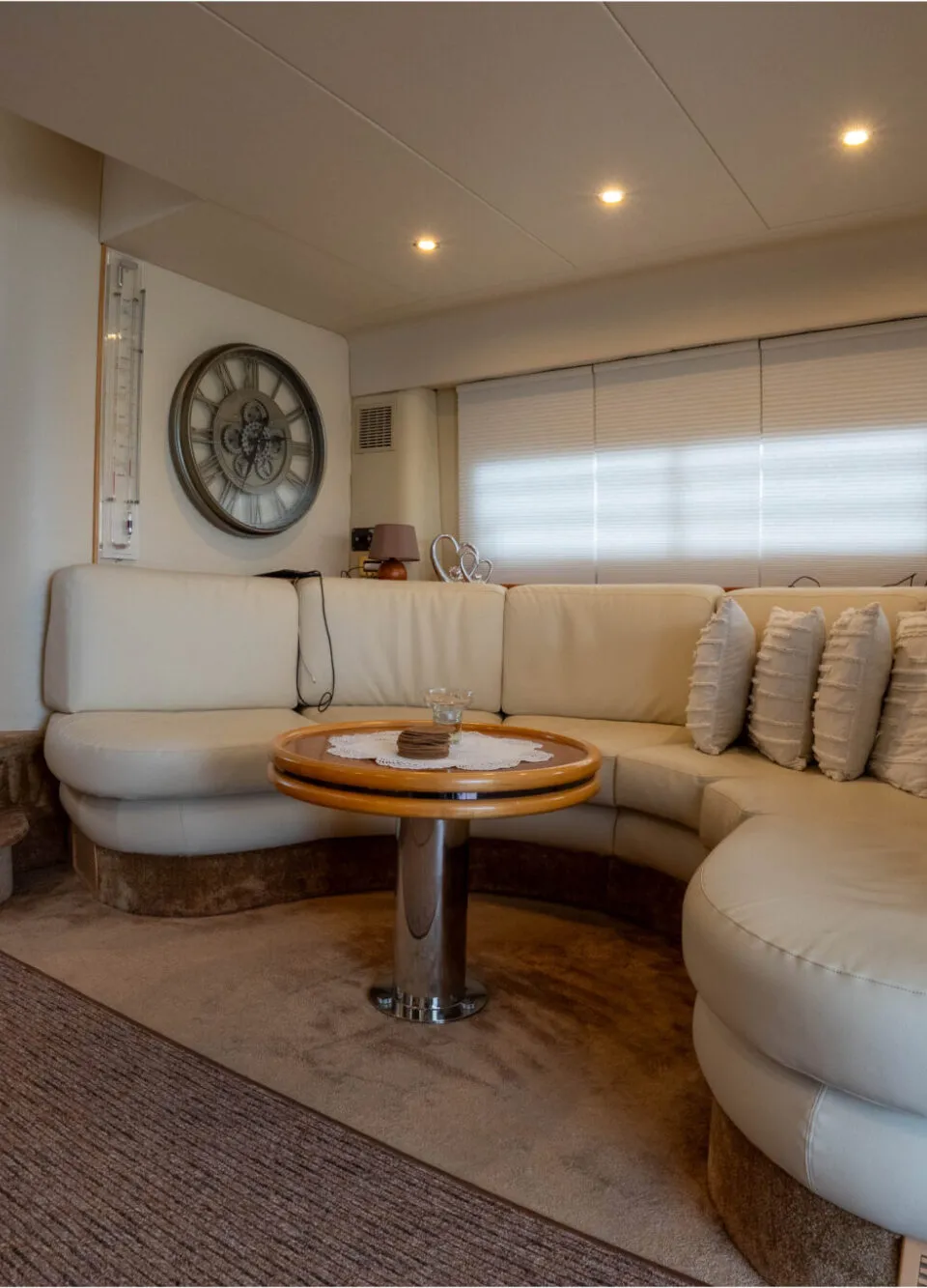 De foto toont een interieur van een boot met een crèmekleurige U-vormige bank, een ronde houten tafel en een tapijt.