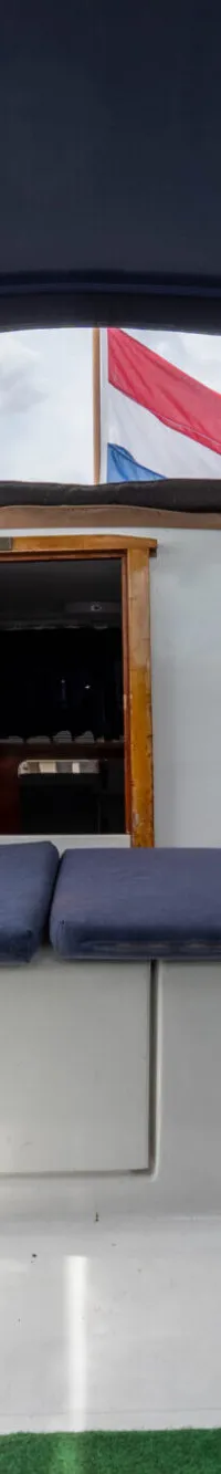 De foto toont het interieur van een boot met een blauwe kap, blauwe kussens en een Nederlandse vlag aan de achterzijde.