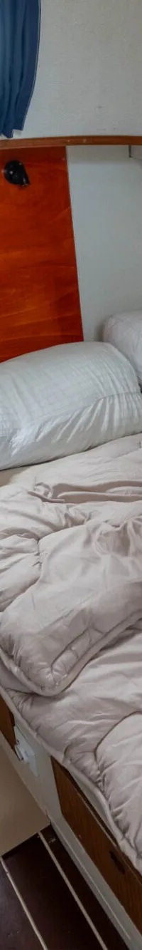 De foto toont een kajuit met een bed, twee kussens, een dekbed, een spiegel en een raam met blauwe gordijnen.