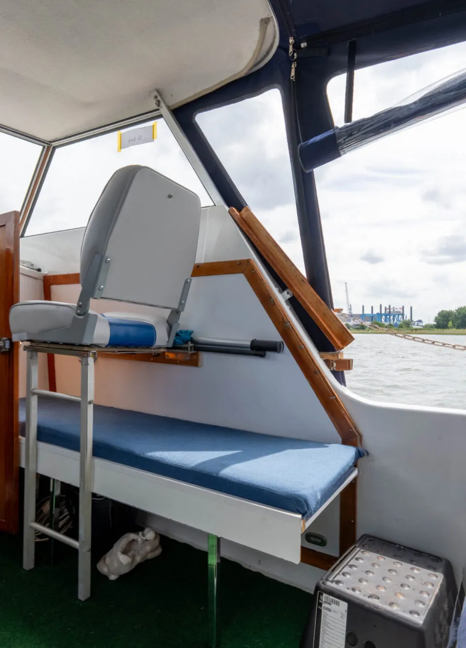 De foto toont het interieur van een boot met een stuurstoel, bankjes en een doorkijk naar de kajuit en het water.