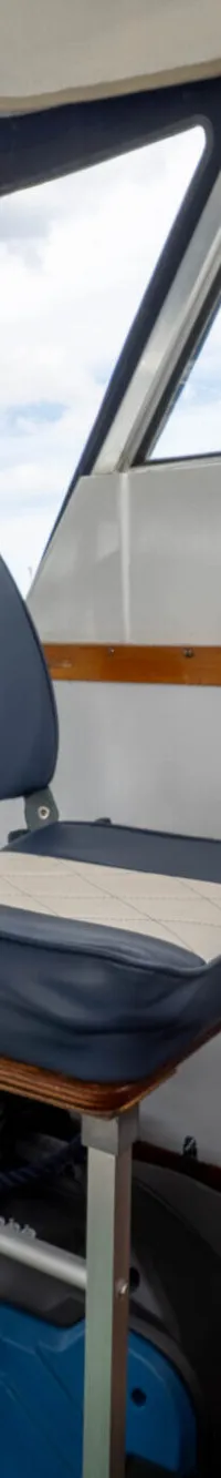 De foto toont het interieur van een boot met een stuurwiel, een blauw-wit gestoffeerde stoel en een bankje.