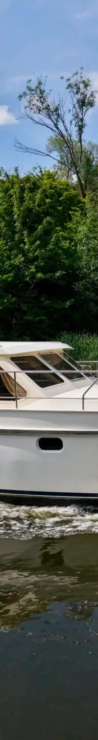 Een witte motorjacht met de naam "Koertje" op de spiegel vaart op het water, omringd door groene bomen onder een blauwe…
