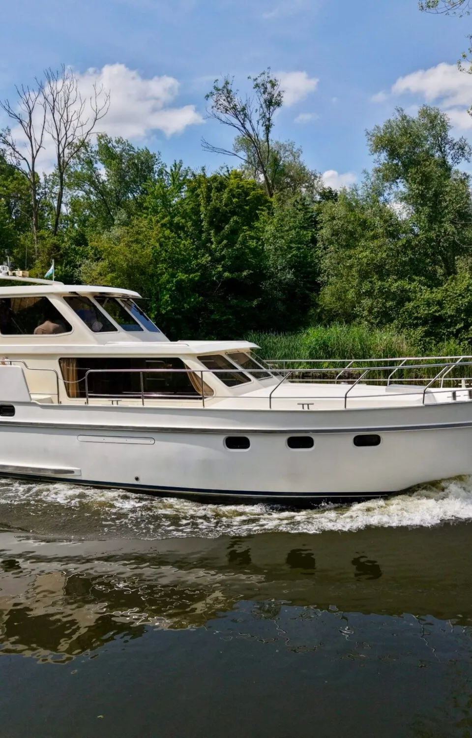 Een witte motorjacht met de naam "Koertje" op de spiegel vaart op het water, omringd door groene bomen onder een blauwe…