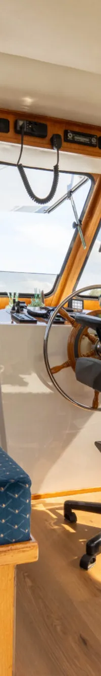 De foto toont een houten interieur van een boot met een stuurwiel, een bureaustoel, een bed en een witte koelkast.