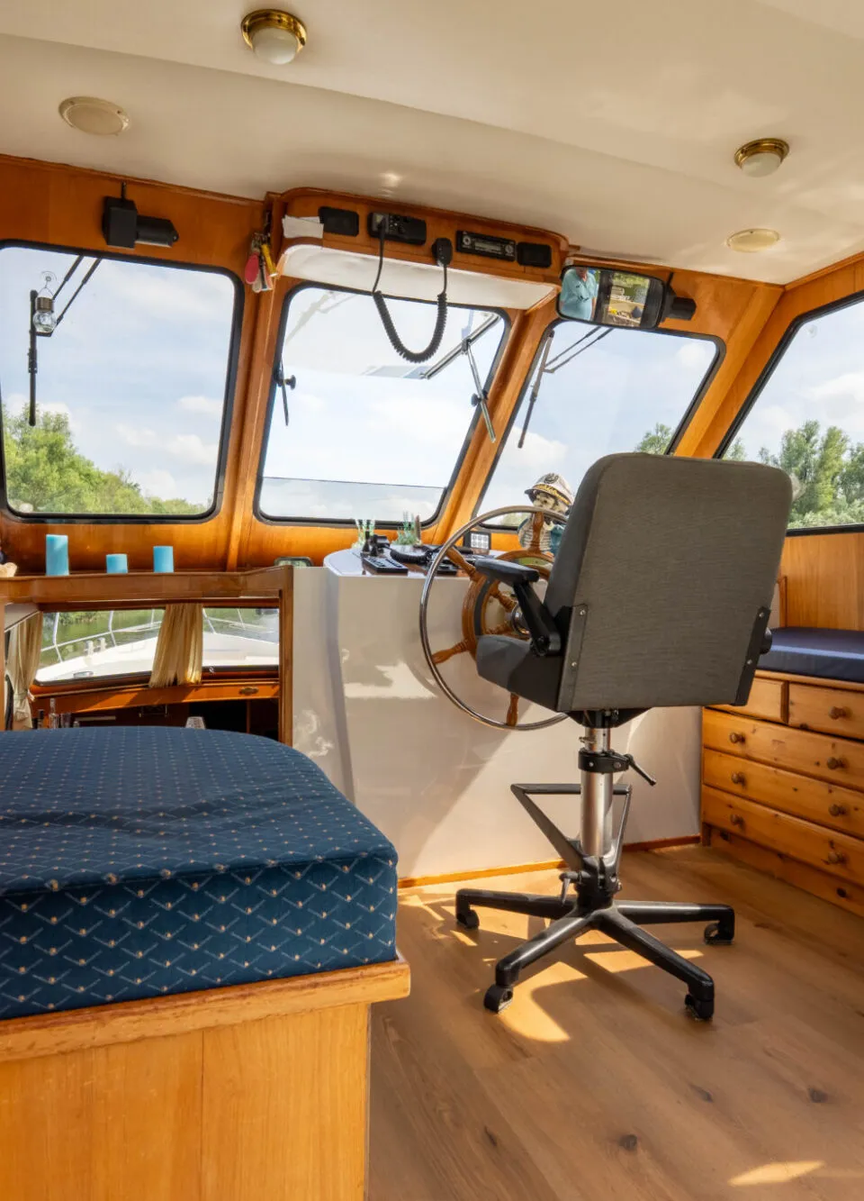 De foto toont een houten interieur van een boot met een stuurwiel, een bureaustoel, een bed en een witte koelkast.