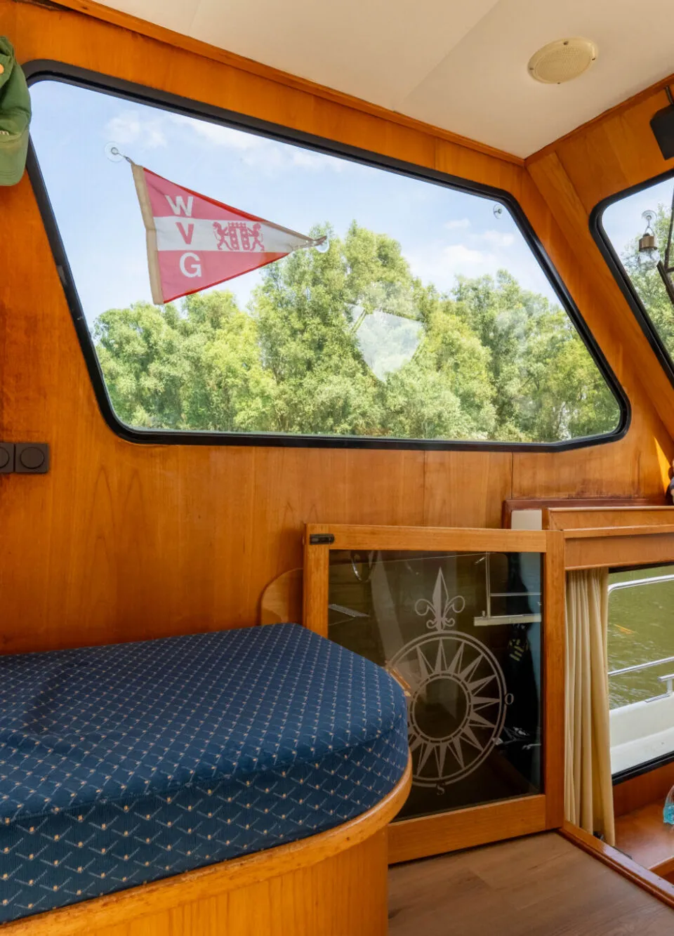 De kajuit toont een houten interieur met ramen, een blauw kussen, een vlag met "W V G" en een kompasroos op een deur.