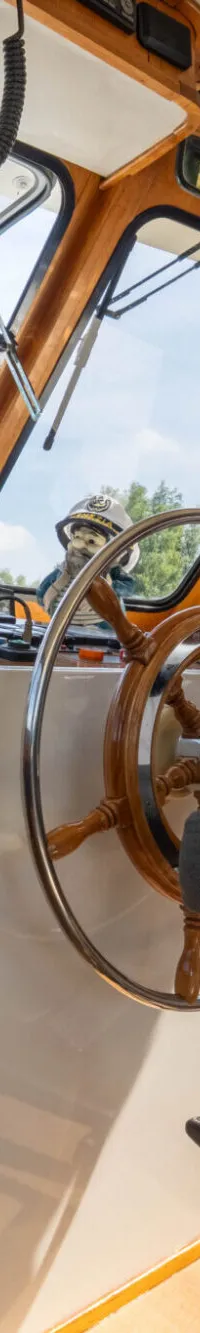 Een stuurhut met een houten stuurwiel, een grijze stuurstoel en een wit dashboard, omringd door ramen en houten betimmering.