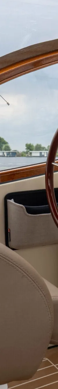 Een beige stuurstoel en een houten stuurwiel zijn zichtbaar in de cockpit van een boot, met uitzicht op water en land.