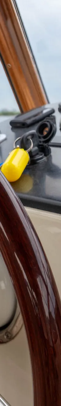 Een houten stuurwiel en bedieningspaneel met diverse knoppen en een joystick zijn zichtbaar in de cockpit van een boot.