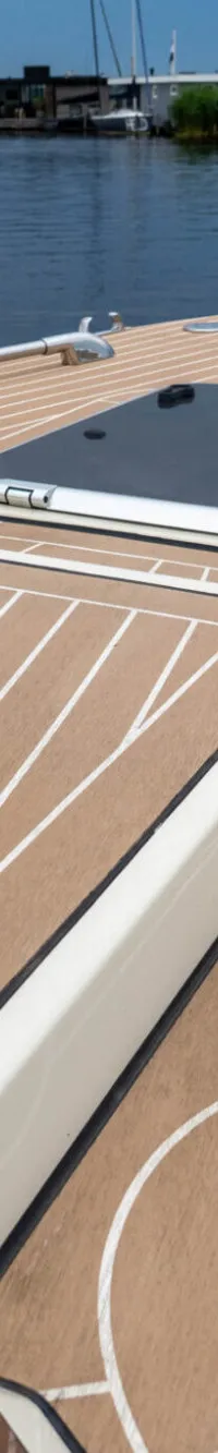 De foto toont het voordek van een boot met een teak-look dek, een luik en een reling, varend op water met woonboten op de…