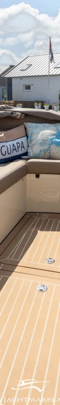 De foto toont het interieur van een boot met beige kussens, een houten vloer en een Nederlandse vlag aan de achterzijde.