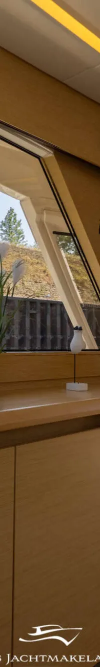 Een interieur van een boot met houten kasten, een raam met uitzicht op een dijk en een open deur naar het dek.
