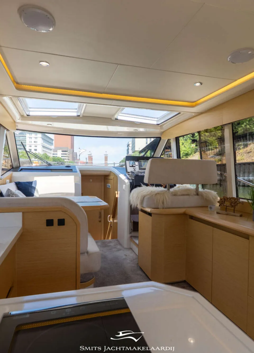 Een interieur van een boot met een keukenblok, zitgedeelte, stuurstand en ramen met uitzicht op water en bebouwing.