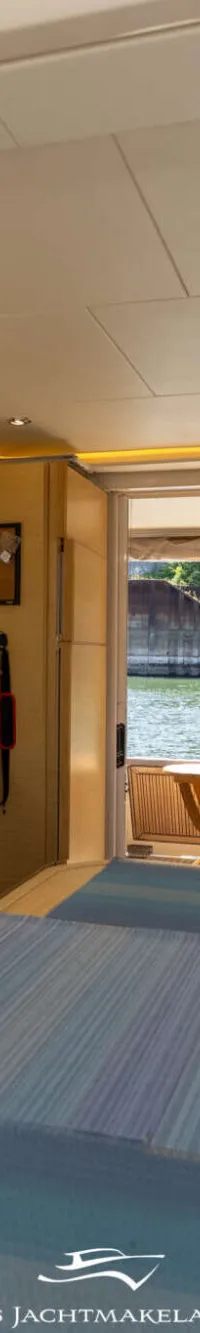 Een interieur van een boot met een houten tafel, banken, een stoel en een keukenblok met uitzicht op water en groen.