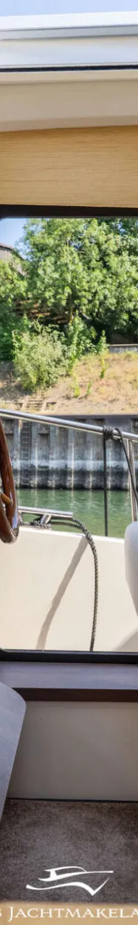 De foto toont het interieur van een boot met een stuurstand, een houten stuurwiel en een bank met uitzicht op water en groen.