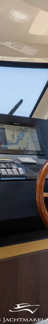 Een stuurstand met een houten stuurwiel, navigatiescherm en diverse knoppen, met uitzicht op een waterweg en gebouwen.