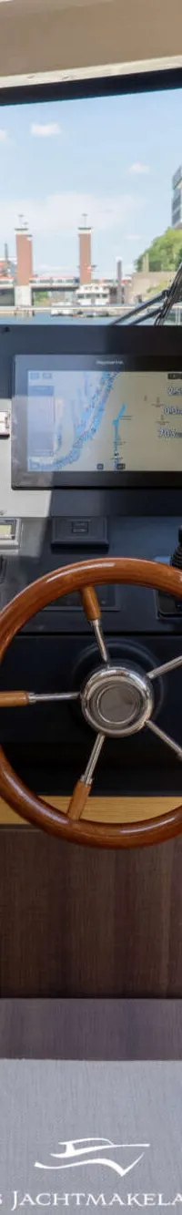 De foto toont het stuurhuis van een boot met een houten stuurwiel, navigatiescherm en bedieningspanelen.