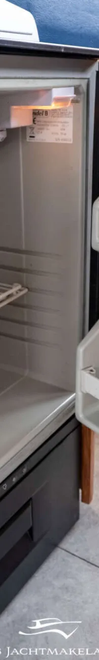 Een openstaande, lege koelkast met een klein vriesvak en een rooster, ingebouwd in een houten interieur.