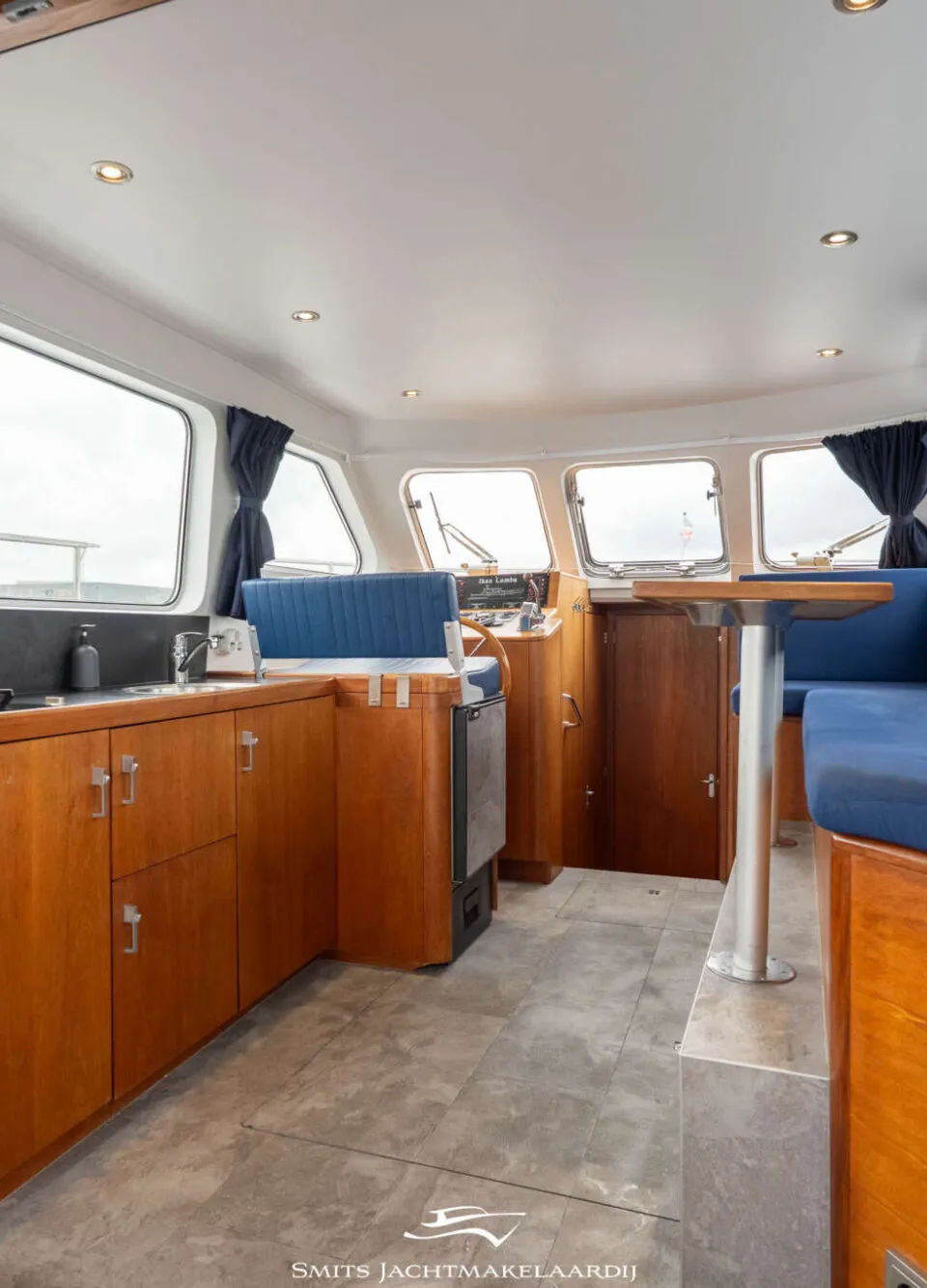 De foto toont het interieur van een boot met een kombuis, zithoek, stuurstand en ramen.