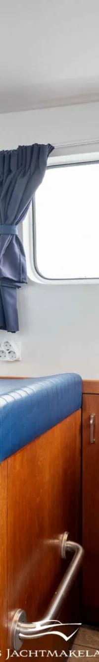 De foto toont het interieur van een boot met een stuurwiel, blauwe bank, houten kasten en een kookplaat.