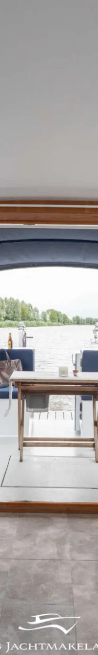 Een interieur van een boot met blauwe banken, houten tafels en een open achterdek met uitzicht op water en land.
