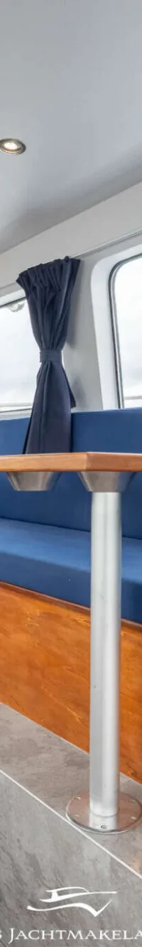 Een interieur van een boot met houten lambrisering, blauwe kussens, een tafel en ramen met uitzicht op groen.