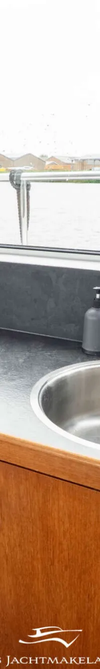 Een kombuis met een aanrecht, spoelbak, kraan, kookplaat en houten kasten, met uitzicht op water en gebouwen.
