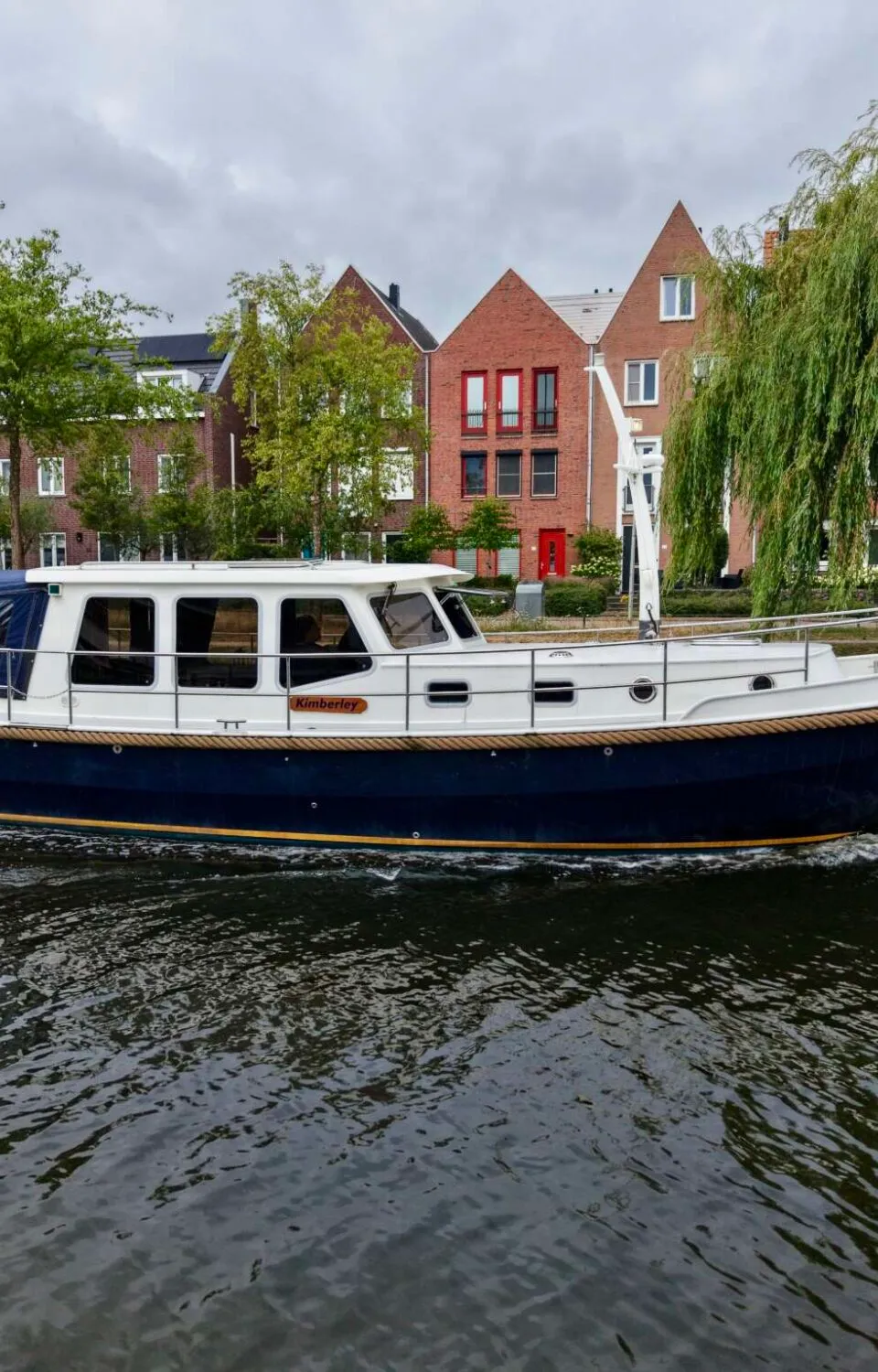 Een blauw-witte motorboot met de naam "Kimberley" vaart op een kanaal langs huizen en bomen. — Brandsma Brandsma 11.00 OK SP