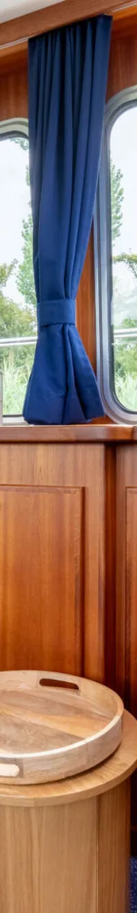 De foto toont een interieur van een boot met houten wanden, ramen met blauwe gordijnen, een rieten stoel en een blauwe stoel.
