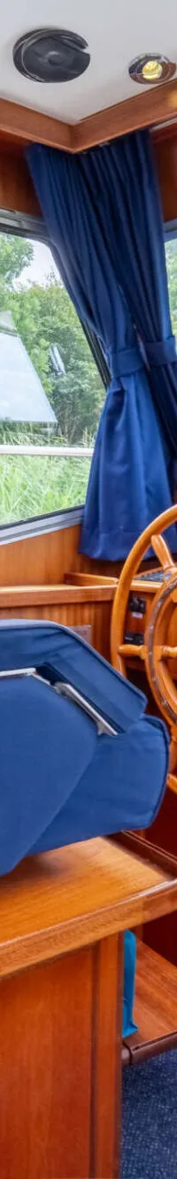 Een interieur van een boot met een houten stuurwiel, een blauwe stoel en ramen met uitzicht op groen.